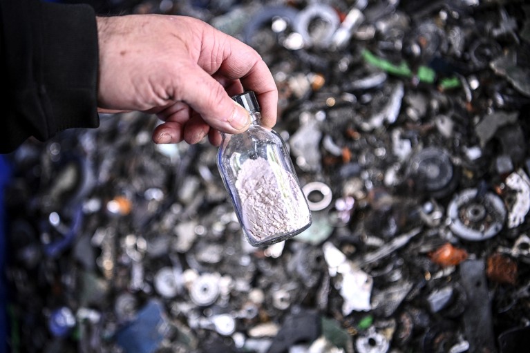 A hand holds a vial filled with rare earths with blurred electronic waste in the background