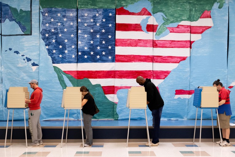 A row of four voters fill out their ballots at a polling station with a painted American flag in the shape of the USA on the wall behind them