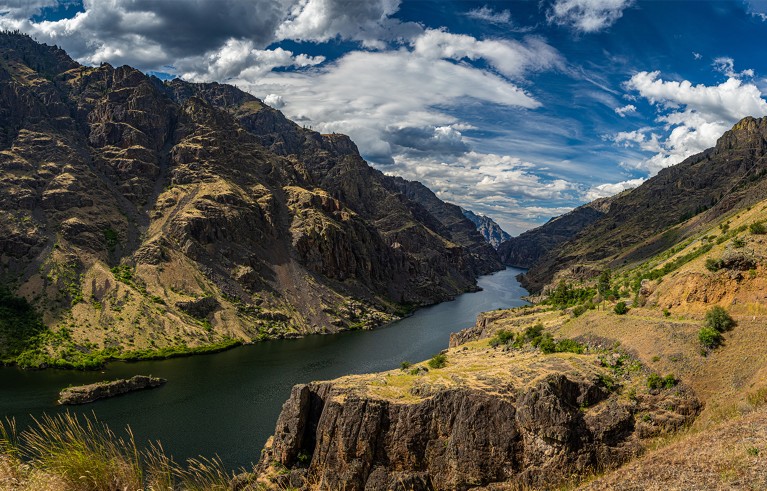 A view of a river through a canyon with mountains around and a blue sky and clouds.