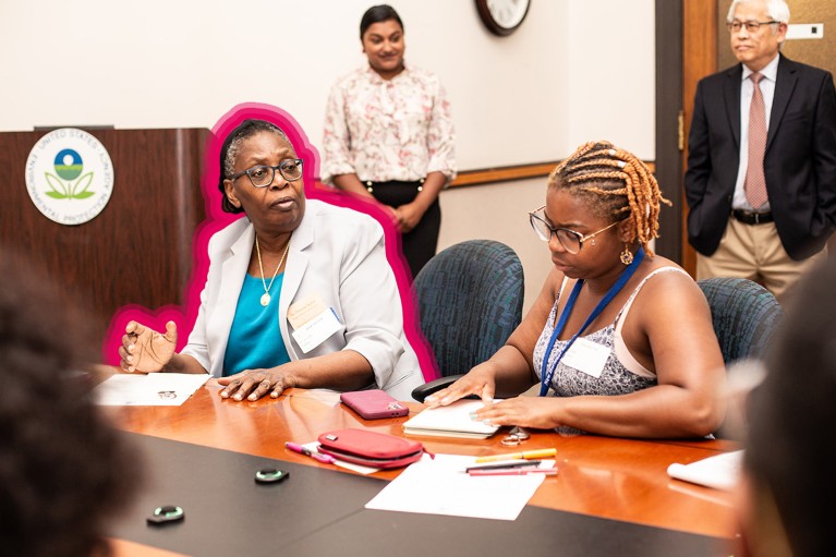Dorceta Taylor, outlined in pink, sits at a table and gestures while talking with other people at a conference