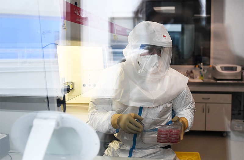 A scientist in a Hazmat suit works with Koch's bacillus in a biomedical laboratory, seen through a glass window.