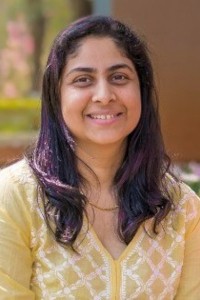 A portrait photograph of Locum Associate Editor Sayali Avachat. Sayali has long dark hair and is wearing a yellow cardigan. She is standing outside and plants are visible in the background.