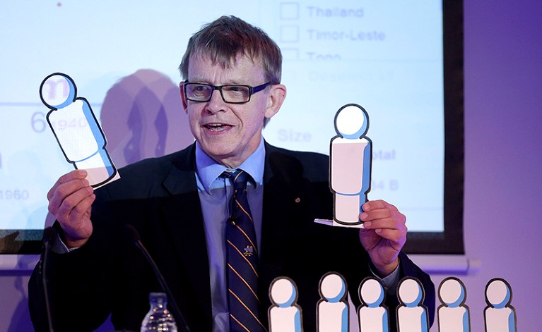 A white man in glasses, wearing a shirt, tie and jacket, holds up cardboard cutouts representing people.