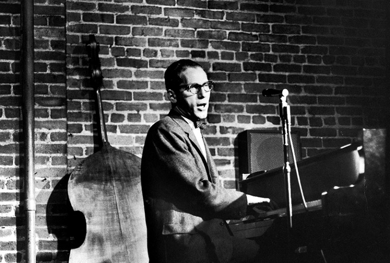 Black and white image of a white man in glasses playing the piano and singing, with a double bass in the background.