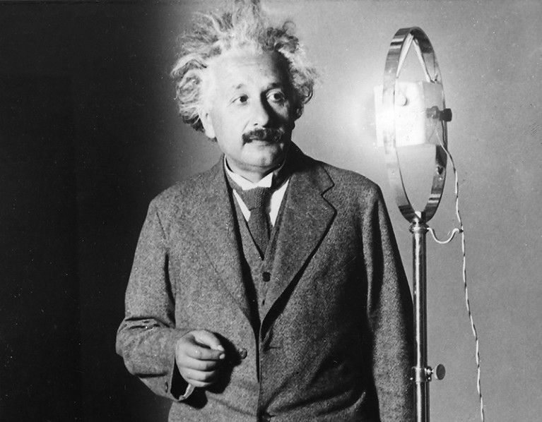 Black and white photo of Albert Einstein, a white man with wild hair and a moustache, standing next to a 1920s microphone.