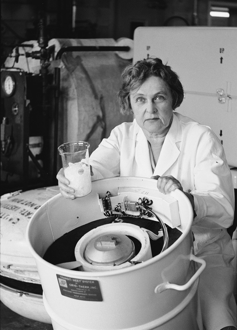 A white woman wearing a lab coat stands next to a large reaction vessel, holding a flask in one hand.