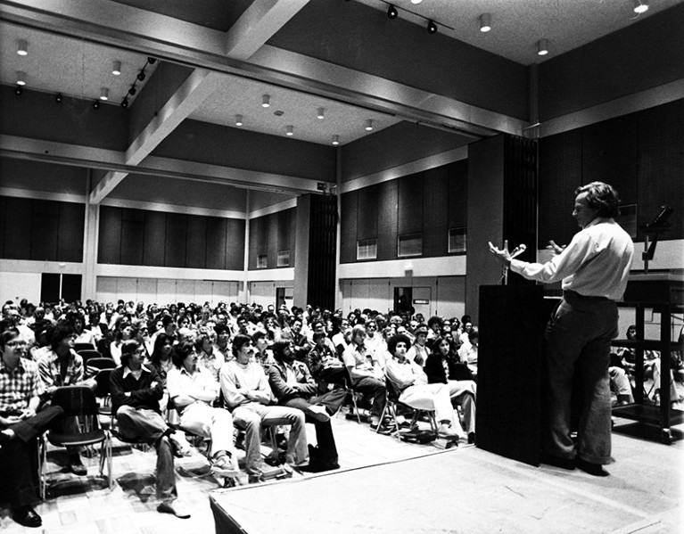 Black and white photo of Richard Feynman standing at a podium with his back to the camera, speaking to a room full of people.