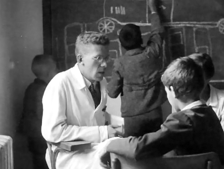 Black and white photo of Hans Asperger, in a lab coat, seated and talking to a boy. Other children write on a blackboard behind.