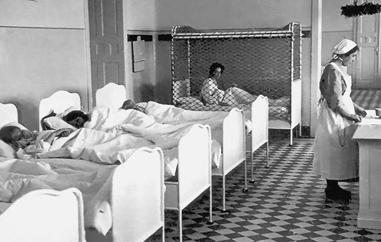 Black and white photo of children lying in a row of hospital beds at Spiegelgrund. A nurse stands facing away from them.