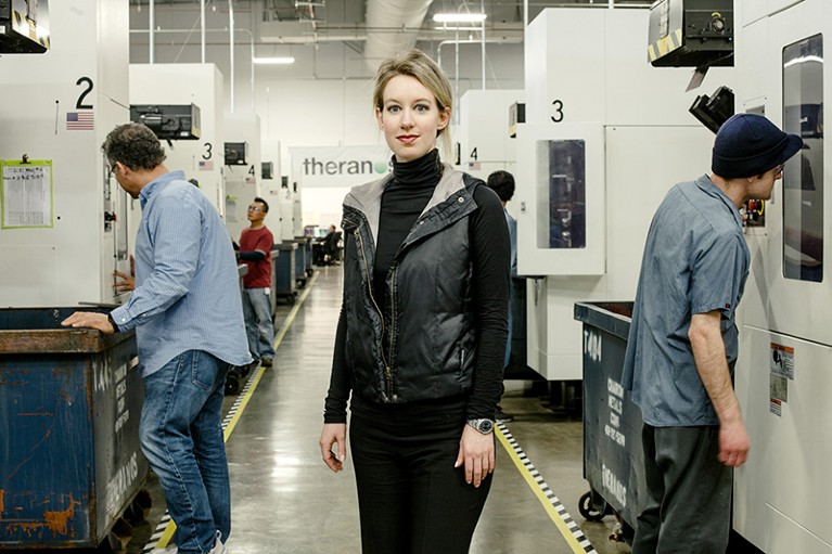 Elizabeth Holmes, a white woman dressed in black, stand between workers and machines in a biotechnology manufacturing facility.
