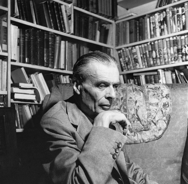 Black and white photograph of Aldous Huxley posing in an armchair in front of bookshelves in Los Angeles.