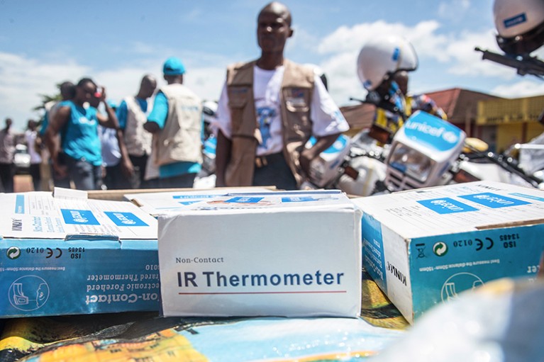 Boxed Infrared thermometers on a table ready to be distributed. UNICEF and UN members stand in the background ready to deploy.