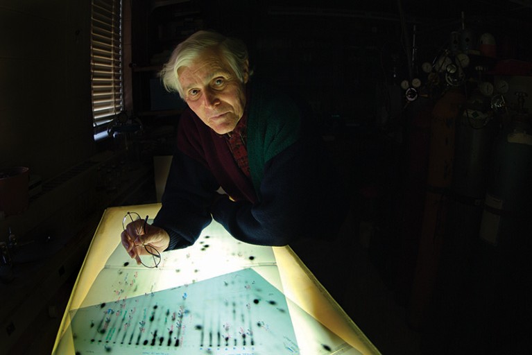Carl Woese, a white man with grey hair and holding a pair of glasses, looks up towards the camera from a lightbox.