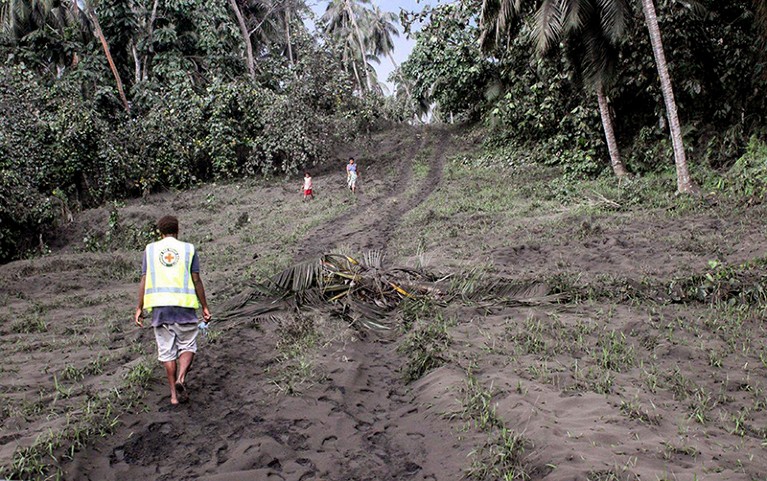 Two distant figures walk towards a Vanuatu Red Cross member (L) wearing high vis across an ash covered forest road on Ambae Isle