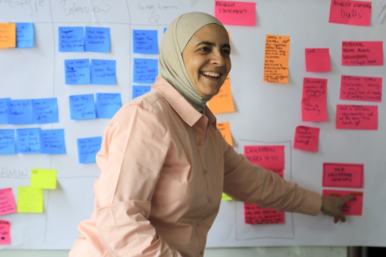 Rana Dajani smiling as she points during a brainstorming session. Behind her is a whiteboard covered with colourful postits