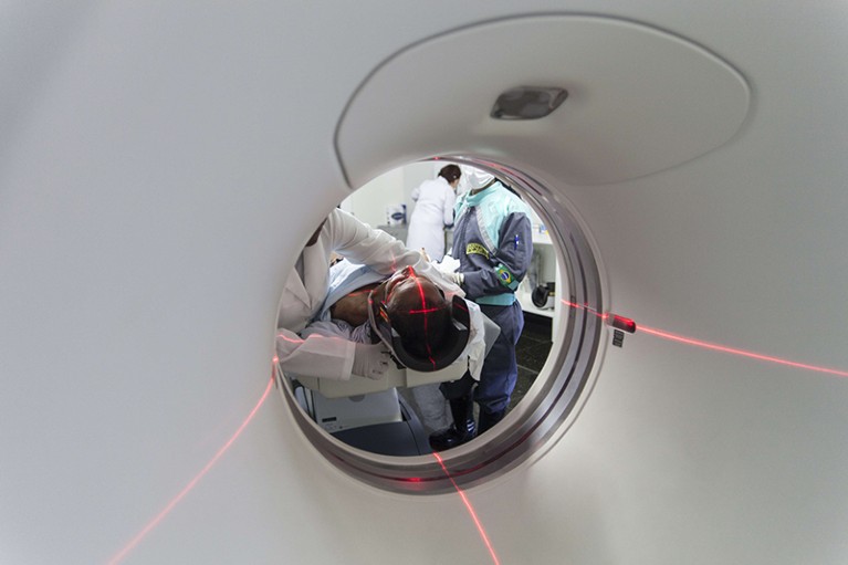 A person entering an MRI machine, guiding lasers are visible in a cross at their head. Hospital staff can be seen in background