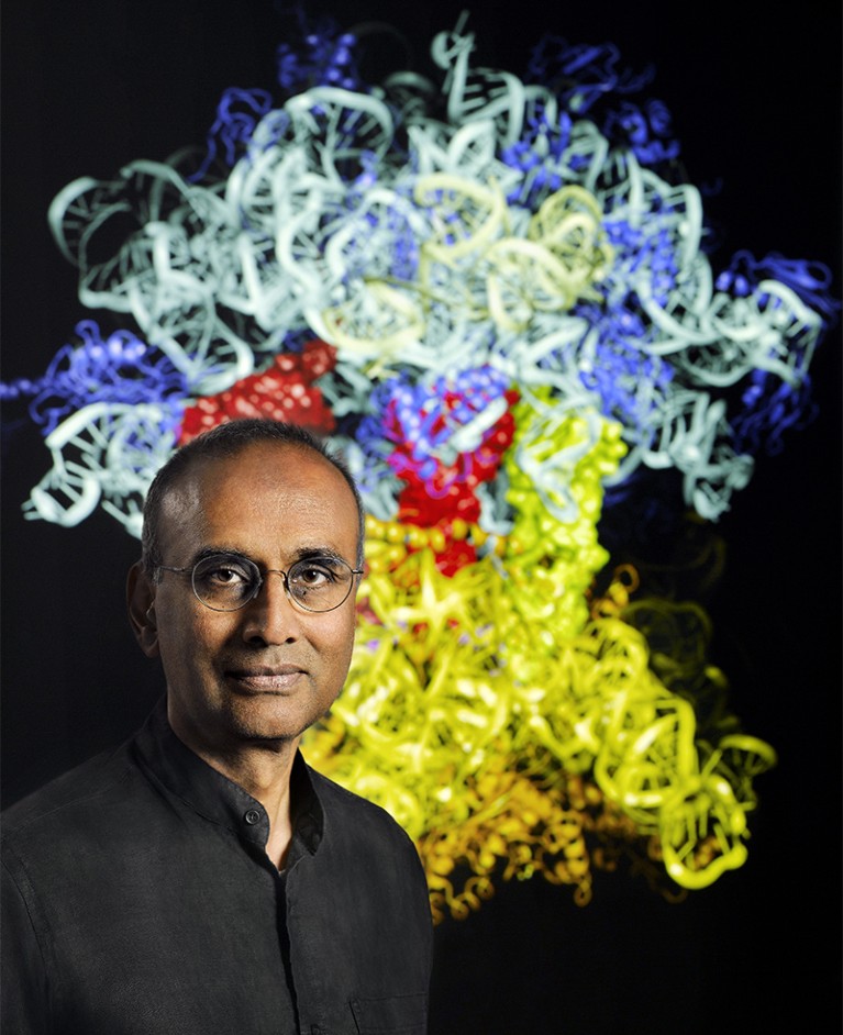 Portrait of Ramakrishnan wearing a black shirt. A ribosome structure can be seen on a black screen behind him