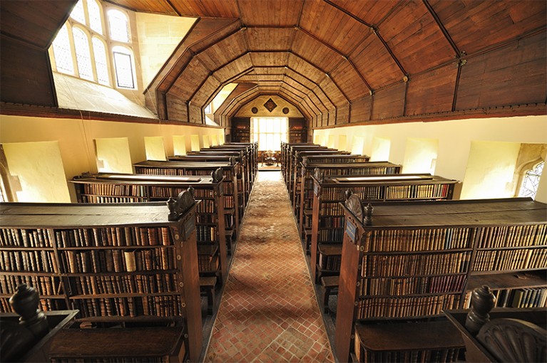 View of Merton College Library. Stacks of books can be seen lined down a tiled hallway.