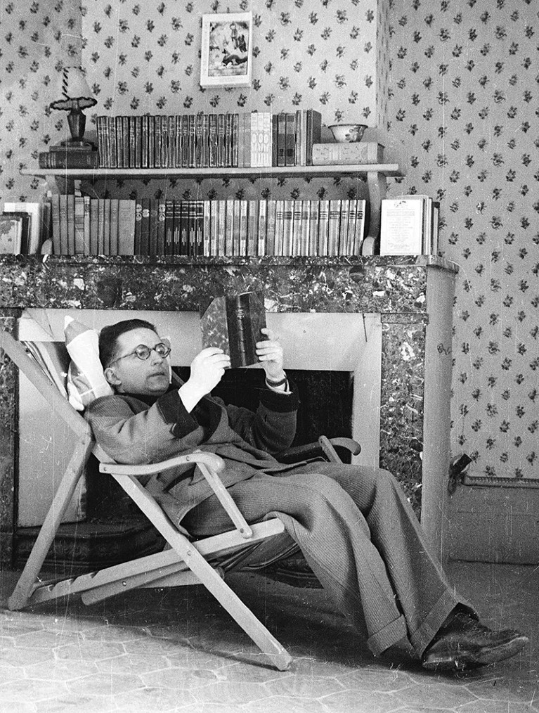 Marian Rejewski reclines in a deck chair reading a book in front of a fireplace