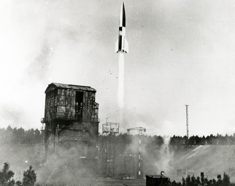 Black and white V2 rocket launching into the air. A small warehouse and forest can be seen in background