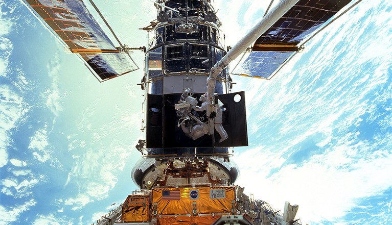 Two astronauts replace gyroscopes on the Hubble telescope during an EVA in 1999. The Earth looms brightly below them.