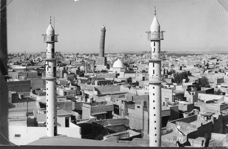 Black and white photo showing three tall minarets and houses in Mosul. The centre minaret is leaning.