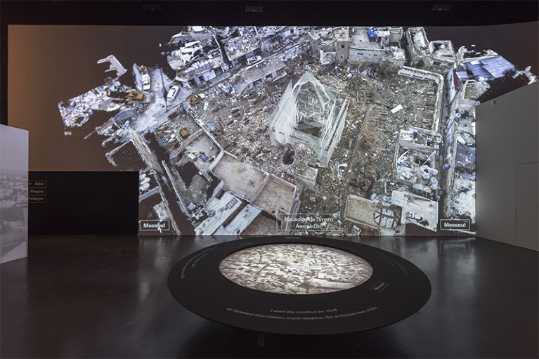 A low table at front showing an aerial view of a city. In the background a large screen shows virtual constructions amid rubble