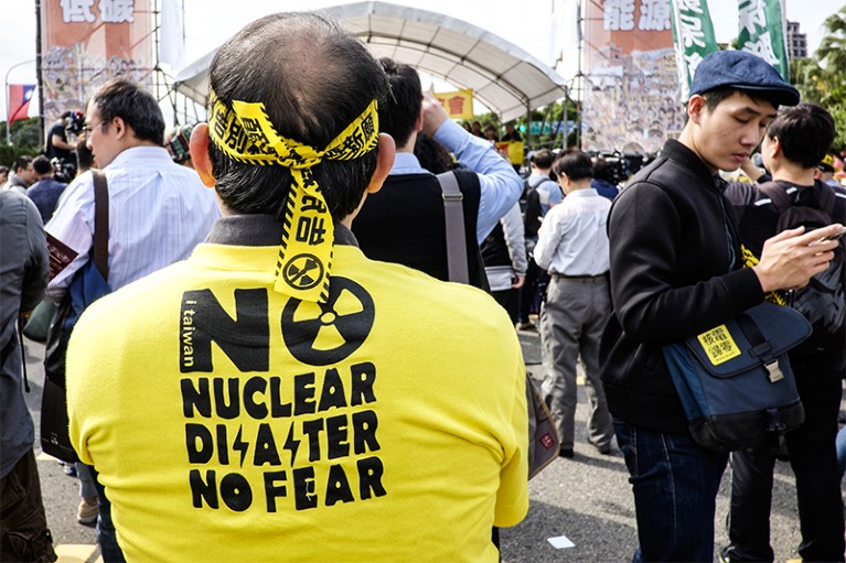 A man in a 'No nuclear no fear' tshirt and with a bandana made from warning tape stands with his back to the camera.