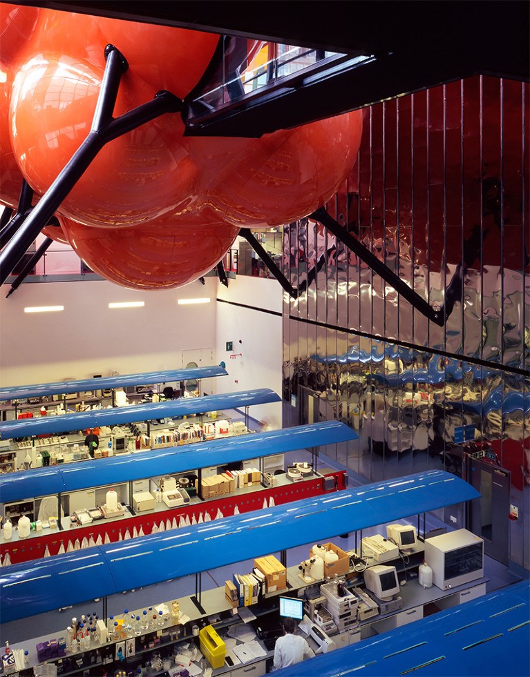 View of the Blizard building with suspended red meeting room structure at top ad colourful open plan lab benches at ground level