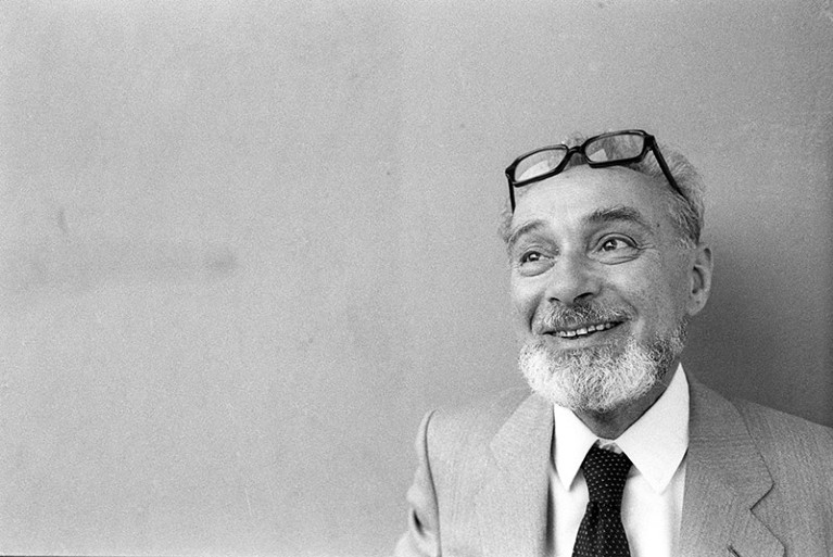 A black and white photograph of Primo Levi, a white man with a greying beard and glasses, standing against a blank wall.