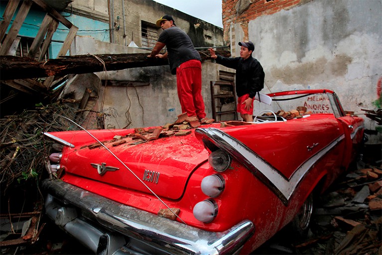 Two men stand on a red car in order to search a destroyed home in Havana