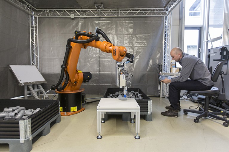 Person sitting on chair uses remote device to control orange robotic arm.