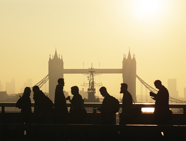 Silhouetted people of various heights walk across London Bridge at sunset.