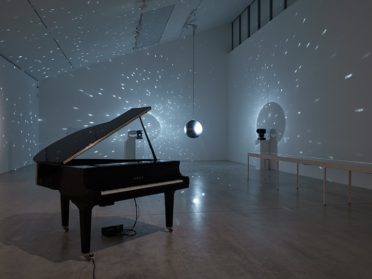 A grand piano stands in a darkened room. Behind it, a mirror ball casts dots of light onto the white walls.