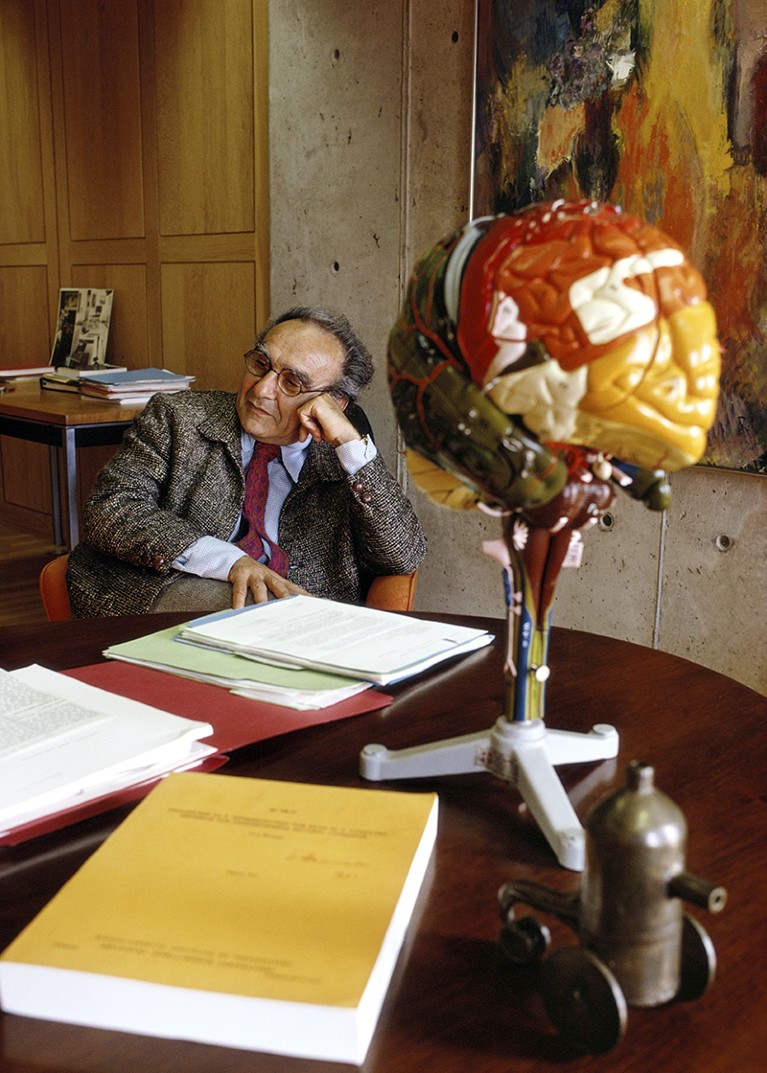 Jacob Bronowski at a desk covered in books. A multicoloured plastic model of a brain on a stand is in the foreground.