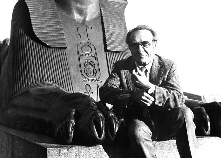 A black and white photograph of Jacob Bronowski sitting between the paws of a stone Egyptian sphinx.