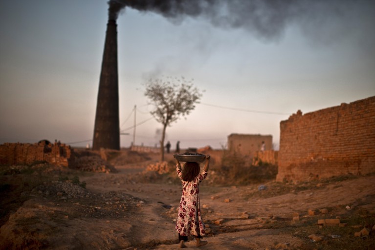 A young girl walks by a smoking chimney carrying a pan of coal on her head, Islamabad, Pakistan.
