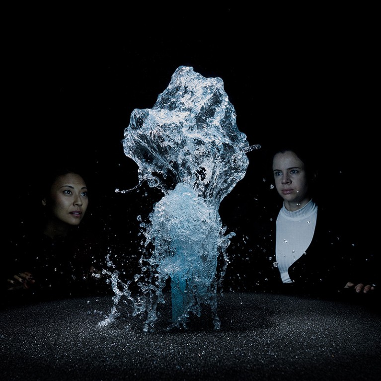Two women look at a spray of blue water bursting between them.