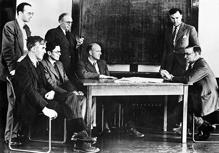 Black and white photo of seven men in suits and ties sitting or standing around a table.
