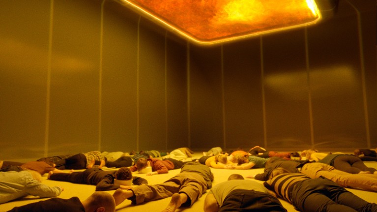 A still from the Swedish film 'Aniara' showing a group of people lying in a small room under a sun panel set in the ceiling