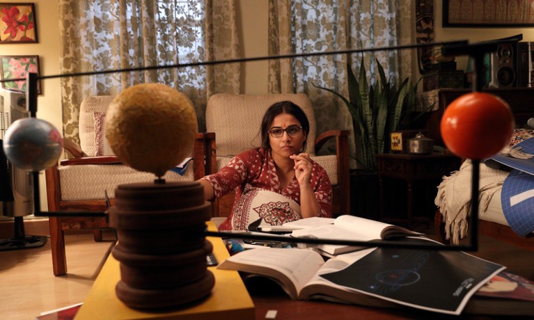 Actress Vidya Balan as project director Tara Shinde - seated at a cluttered desk in front of a solar system model.