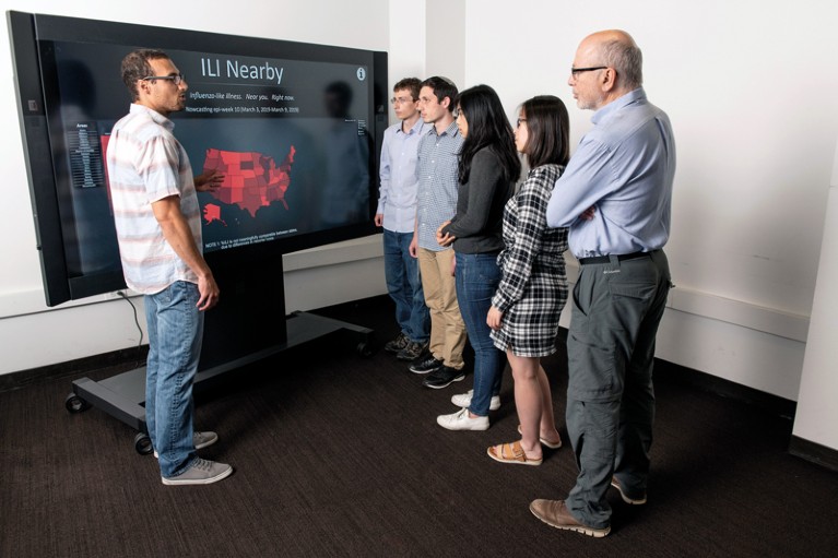 A person stands on the left in front of a screen giving a presentation to five people