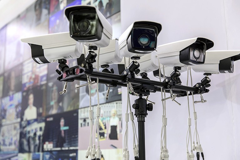 Seven surveillance cameras on a single pole, in front of a screen showing many images.