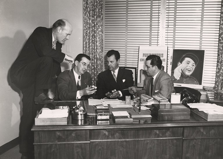 Four men behind a desk discuss cigarette advertisements in an office, 1950s
