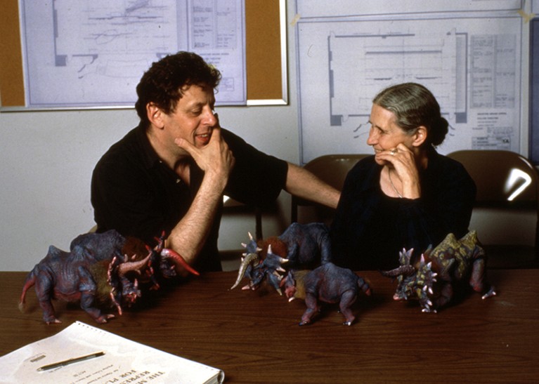 Philip Glass (left) and Doris Lessing (right) in 1988. They are sitting at a table covered in model dinosaurs.