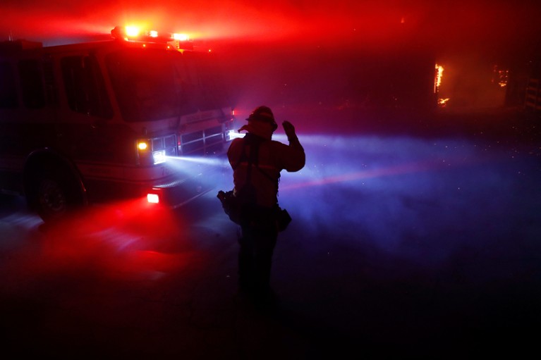 A firefighter walks through thick smoke illuminated by fire engine lights and embers