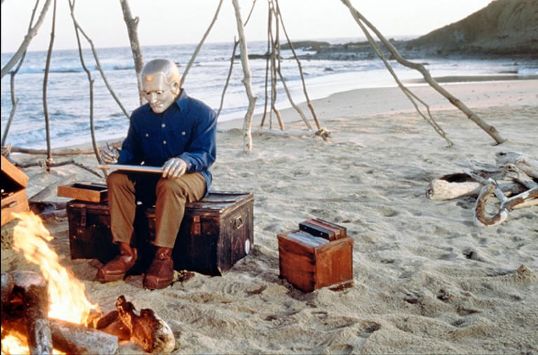 A robot wearing a coat, trousers and shoes sits on a trunk on a beach, next to a campfire.