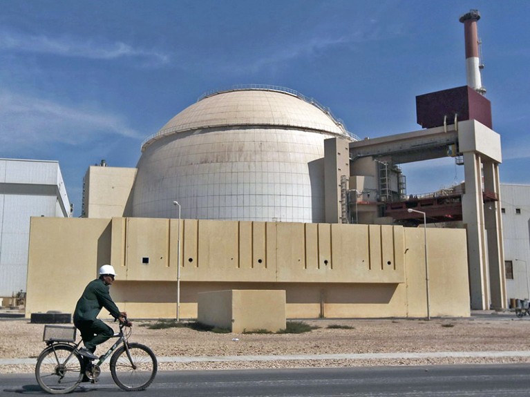 A worker rides a bicycle in front of a reactor building, a white dome behind a concrete wall.