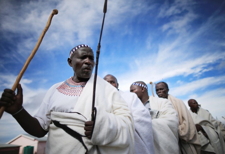 Xhosa people standing in a line, wearing white robes and carrying sticks.