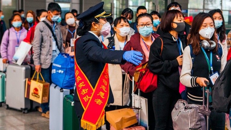 People in face masks queue with luggage, directed by an official in a face mask and gloves.